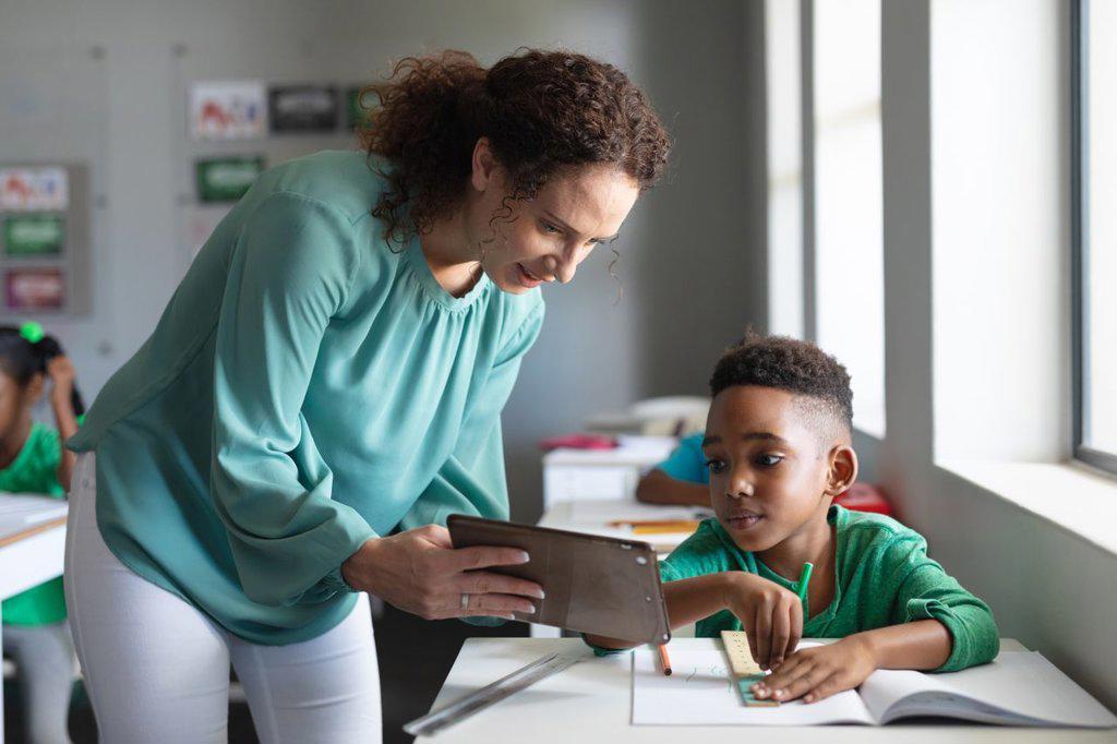 Caucasian young female teacher showing digital tablet to african american elementary boy in class. unaltered, education, childhood, occupation, technology, teaching and school concept.