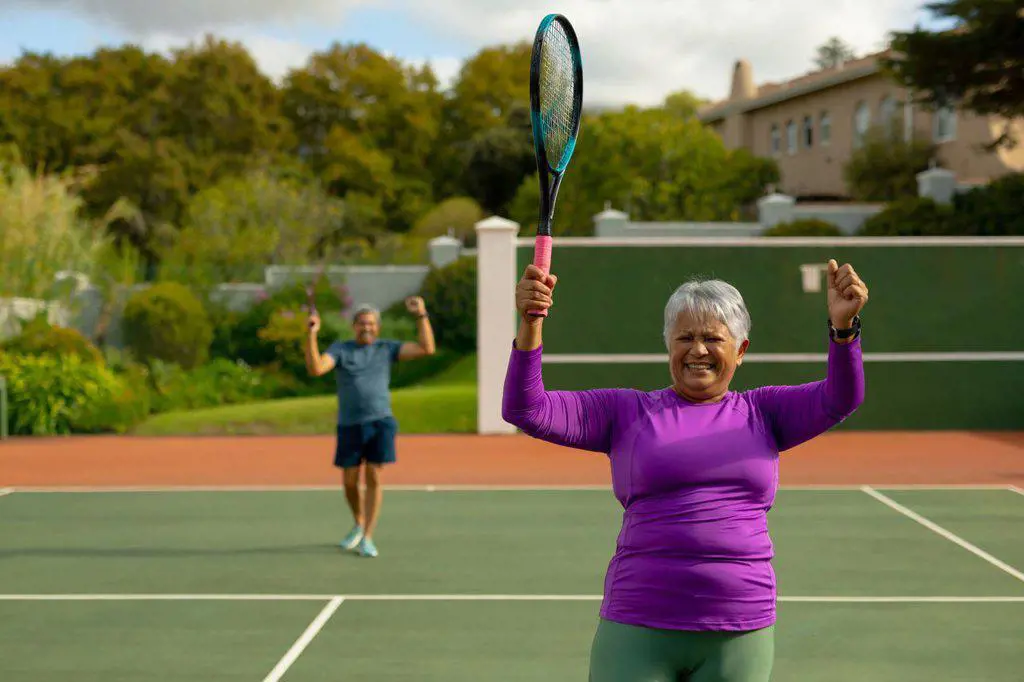 Happy biracial senior woman holding racket and gesturing at tennis court with man in background. achievement, winning, unaltered, sport, competition, retirement, healthy and active lifestyle.