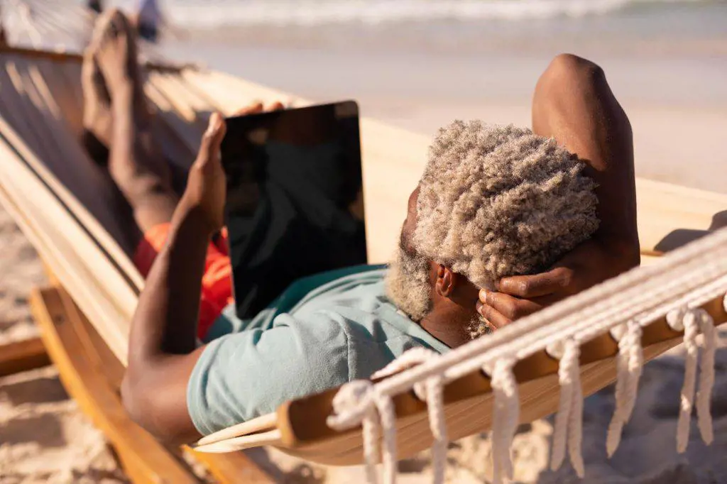 African american senior man with gray hair using digital tablet while lying on hammock at beach. summer, copy space, technology, relaxation, unaltered, lifestyle, retirement, enjoyment, holiday.