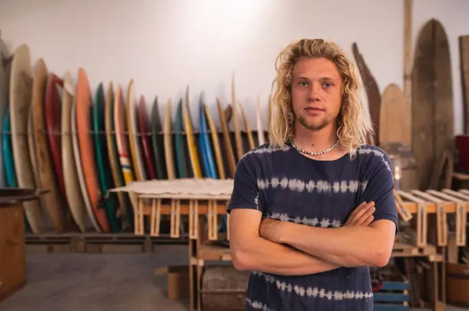 Portrait of a Caucasian male surfboard maker in his studio, with surfboards in a rack in the background, standing with his arms crossed and looking straight to camera. 