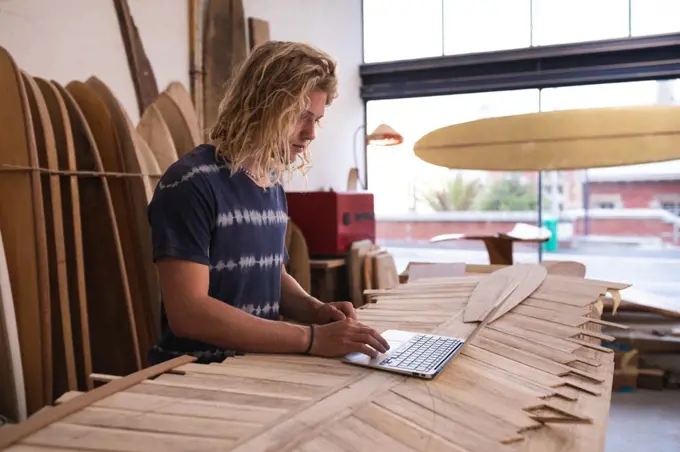 Caucasian male surfboard maker in his studio, working on a project using his laptop, with surfboards in a rack in the background. 