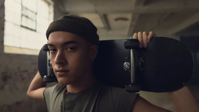 Close up view of a young Hispanic-American man with piercings wearing a dark grey shirt and beanie while holding a skateboard over shoulder and looking intently at the camera while standing inside an empty warehouse
