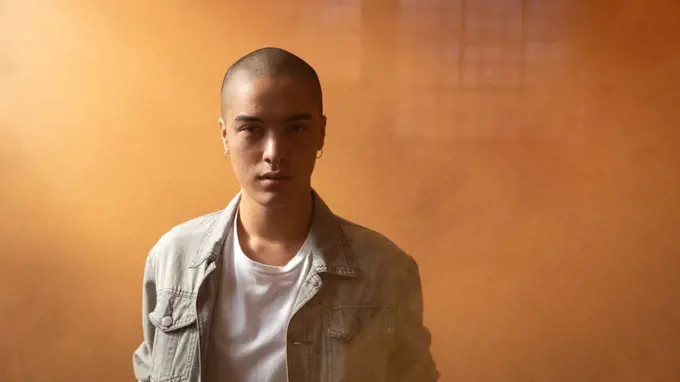 Front view of a young Hispanic-American man wearing a grey leather jacket over a white shirt looking intently at the camera inside an empty warehouse with orange smoke