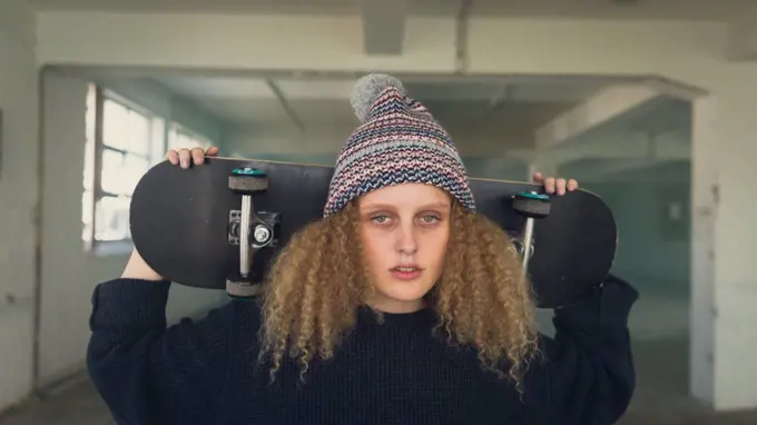 Front view of a young Caucasian woman with curly hair wearing a black long sleeves and beanie while holding a skateboard overshoulder and looking intently at the camera inside an empty warehouse