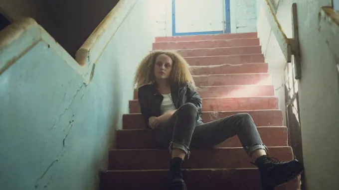 Low angle view of a young Caucasian woman with curly hair wearing leather jacket sitting on the stairs while looking intently at the camera