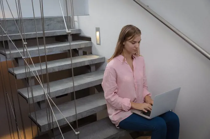 Side view of a young Caucasian woman wearing a pink shirt, sitting on a staircase in an apartment using a laptop computer. She is relaxing and practicing selfcare.