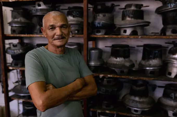 Portrait of a smiling senior mixed race man with arms crossed, standing in front of shelves of traditional equipment in the workshop at a hat factory