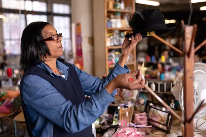 Side view close up of a middle aged mixed race woman inspecting and adding the finishing touches to a hat on a stand in the workshop at a hat factory, with materials visible in the background