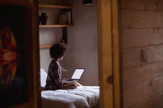 Side view of a young mixed race woman using a laptop computer sitting on her bed at home, seen reflected in the mirror in her room