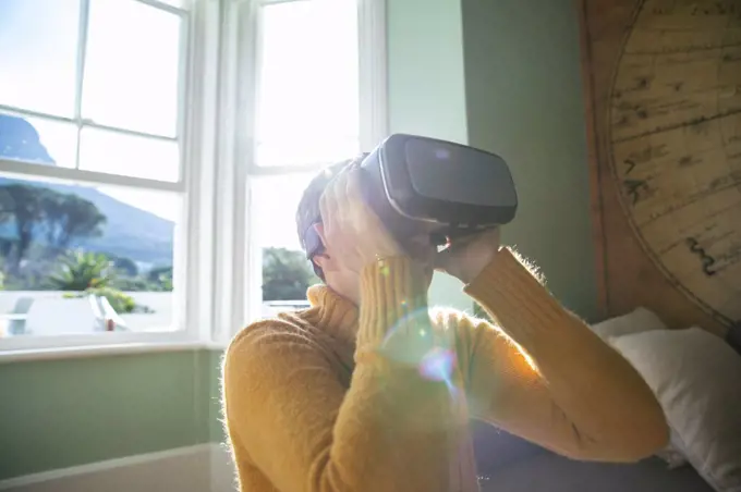 Side view close up of a mature Caucasian woman with short grey hair sitting at home in her living room wearing a VR headset, backlit by sunlight from a window behind her
