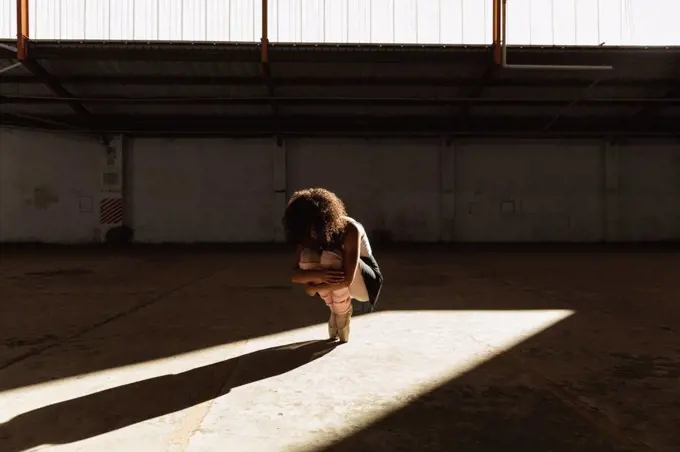 Front view of a young mixed race female ballet dancer wearing pointe shoes squatting down holding her knees balanced on her toes in shaft of sunlight while dancing in an empty room at an abandoned warehouse
