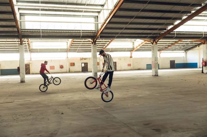Side view of two young Caucasian men facing opposite directions balancing on the back wheels of their BMX bikes while practicing tricks in an abandoned warehouse, the rider in the foreground is jumping off the ground