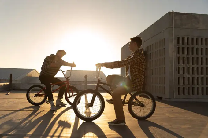 Side view of two young Caucasian men sitting on BMX bikes talking on the rooftop of an abandoned warehouse, backlit by the setting sun, with buildings in the background