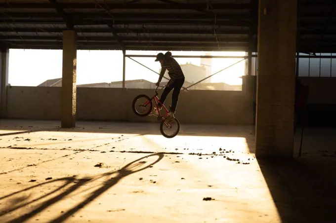 Side view of a young Caucasian man jumping on a BMX bike while practicing tricks in an abandoned warehouse, backlit by sunlight
