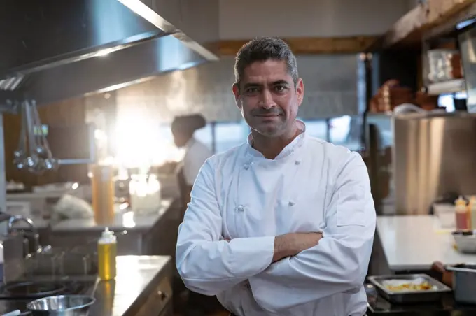 Portrait close up of a smiling middle aged Caucasian male chef wearing whites standing with arms crossed in a restaurant kitchen