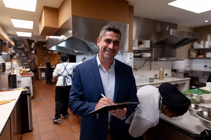 Portrait close up of a middle aged Caucasian male restaurant manager writing on a clipboard in a busy restaurant kitchen, while kitchen staff work in the background