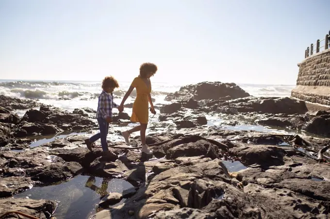 Side view of a young mixed race woman and her pre-teen son enjoying time together by the sea, holding hands and walking on a sunny day