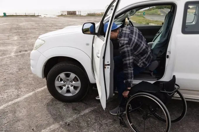 Side view of a young Caucasian man getting out of a car into a wheelchair in a car park by the sea