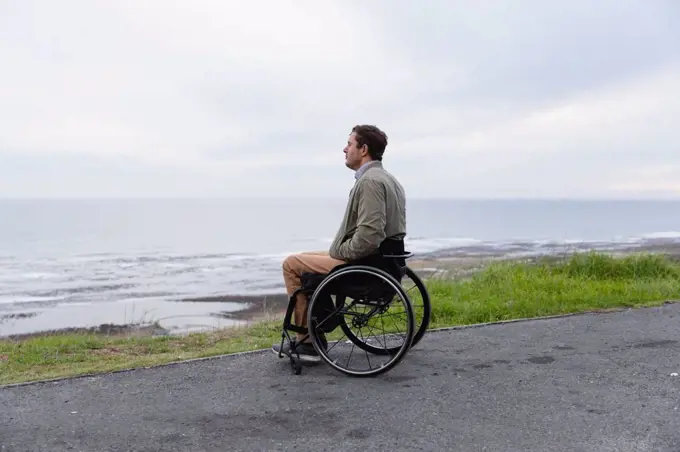 Side view of a young Caucasian man in a wheelchair enjoying a day out by the sea, enjoying the view