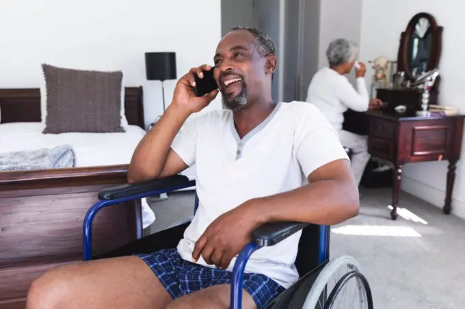A senior retired African American couple at home in their bedroom, the man sitting in a wheelchair in his underwear talking on a smartphone and smiling, the woman sitting at her dressing table in the background, self isolating during coronavirus covid19 pandemic