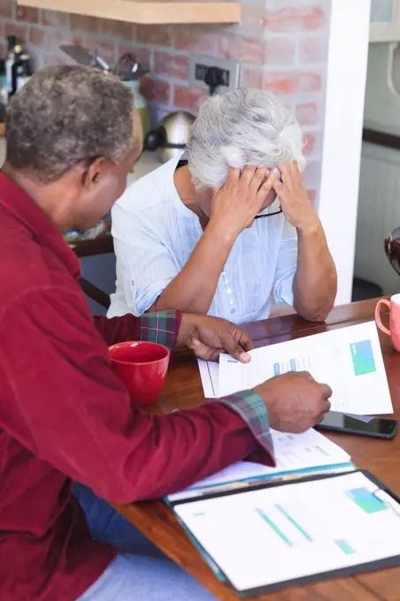 A senior retired African American couple sitting at a table in their dining room drinking coffee, looking at paperwork and discussing their finances, the woman with her head in her hands, at home together isolating during coronavirus covid19 pandemic