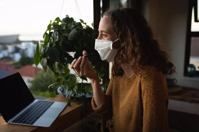 Caucasian woman spending time at home self isolating and social distancing in quarantine lockdown during coronavirus covid 19 epidemic, wearing a face mask against covid19 coronavirus, standing by a window, talking on her smartphone and using a laptop computer.