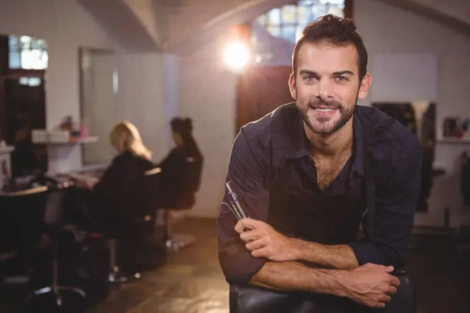 Portrait of smiling male hairdresser leaning on chair in salon