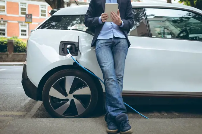 Low section of businessman using digital tablet while charging electric car at charging station