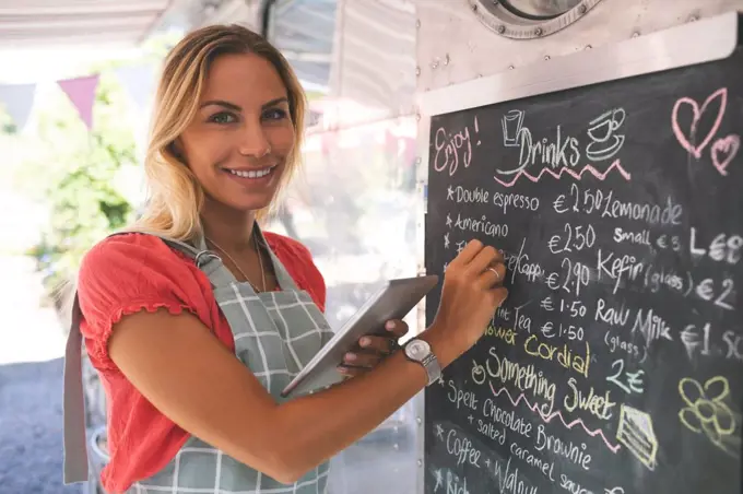 Portrait of female waitress writing menu on menu board while using digital tablet