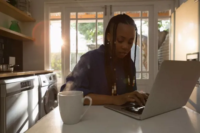 Woman using laptop in kitchen while having coffee on table at home