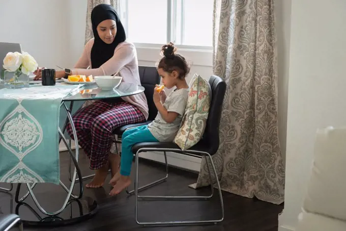Side view of mixed race Mother wearing hijab talking with daughter while daughter eating orange fruit while sitting at table at home