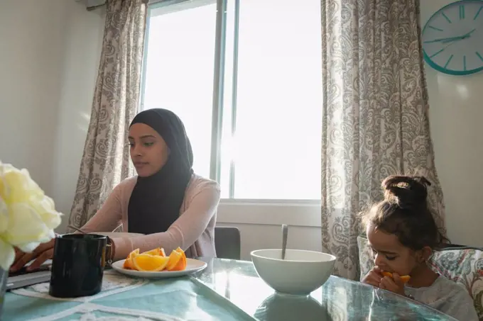 Low angle view of mixed race mother wearing hijab using laptop while daughter eating fruit at home 