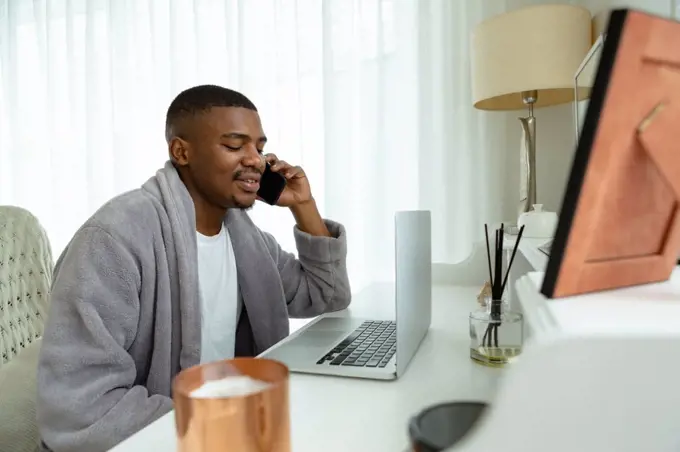 Side view of African-american man talking on mobile phone while working on laptop at desk in bedroom at comfortable home. Authentic home lifestyle setting with young African American male