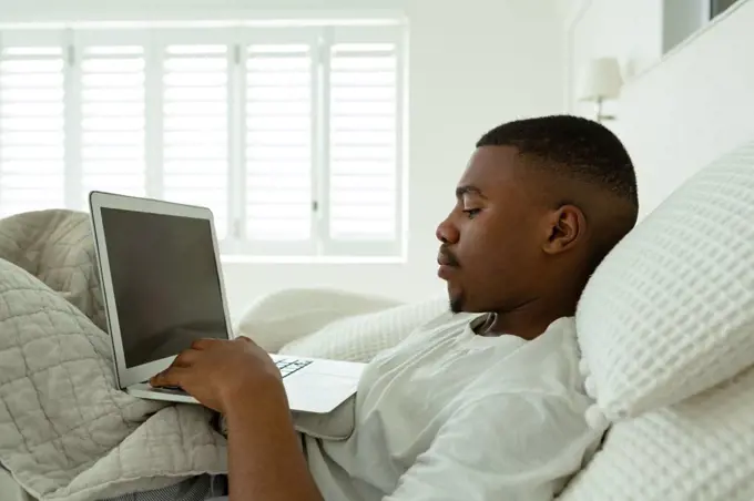 Side view of African-american man using laptop while lying on bed in bedroom at comfortable home. Authentic home lifestyle setting with young African American male