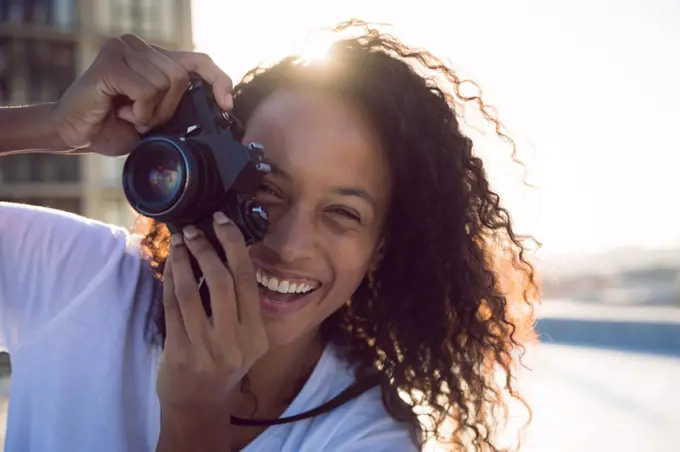 Front view of a young African-American woman smiling while holding a camera and standing on a rooftop with a view of a building and the sunlight. Bright modern gym with fit healthy people working out and training