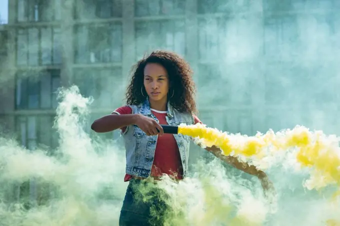Front view of a young African-American woman wearing a denim vest holding a smoke maker producing yellow smoke on a rooftop with a view of a building and sunlight
