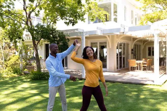Self isolation in quarantine lock down. front view of an african american man and a mixed race woman outside their house in the garden on a sunny day, having fun holding hands and dancing together