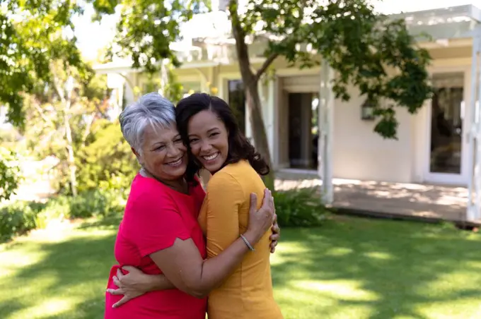 Self isolation in quarantine lock down. side view of a mixed race woman and her senior mother standing outside their house in the garden on a sunny day, embracing, their heads together and both smiling to camera