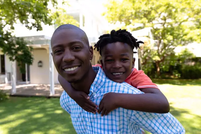 Self isolation in quarantine lock down. portrait of an african american man outside outside his house in the garden on a sunny day, piggybacking his young son, both smiling to camera