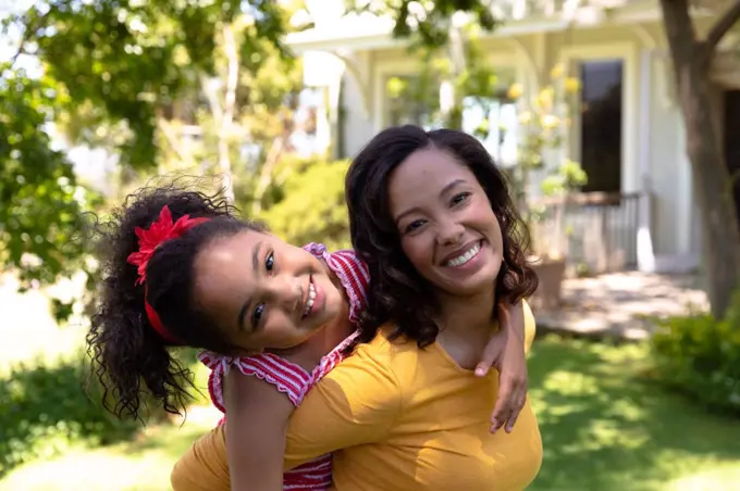 Self isolation in quarantine lock down. side view of a mixed race woman outside her house in the garden on a sunny day, piggybacking her young daughter, both smiling to camera