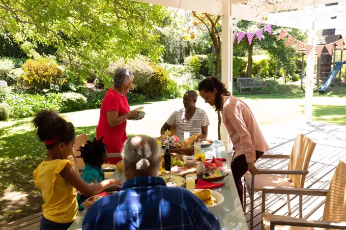 Side view of a multi-ethnic, multi-generation family serving food and sitting down at a table for a meal together outside on a patio in the sun