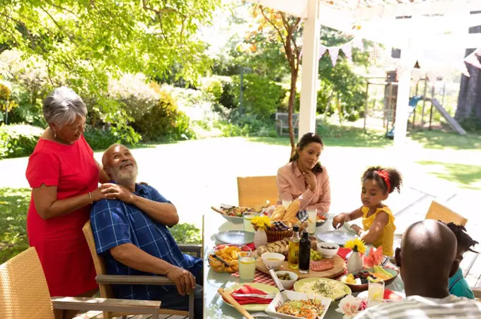 Side view of a multi-ethnic, multi-generation family sitting at a table for a meal together outside on a patio in the sun, the grandparents embracing, the grandmother standing behind the grandfather