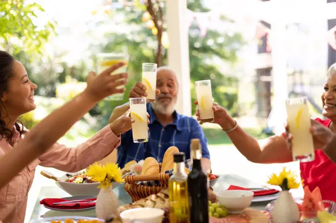 Side view close up of a multi-ethnic, multi-generation family sitting at a table for a meal together outside on a patio in the sun, raising glasses of lemonade and making a toast