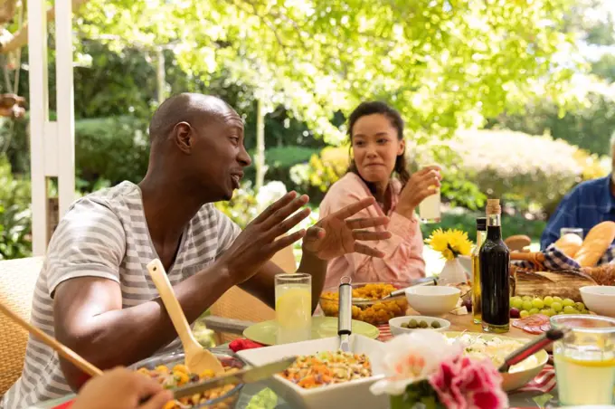 Side view of an African American man talking and gesturing and a mixed race woman listening, sitting and smiling at a table during a with family meal outside on a patio in the sun