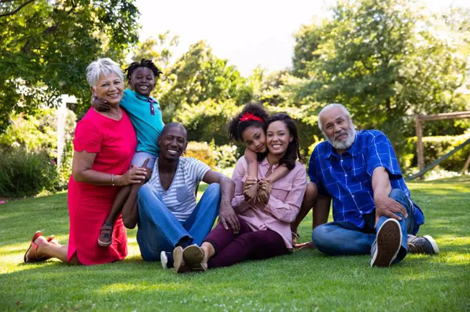 Front view of a multi-ethnic, multi-generation family outside in the garden, sitting on the grass together embracing, looking to camera and smiling