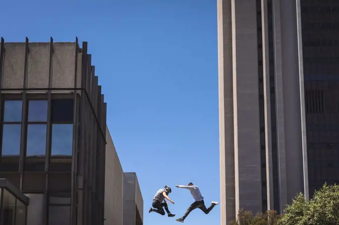 Side view of two Caucasian men practicing parkour by the building in a city on a sunny day, jumping up between modern buildings.
