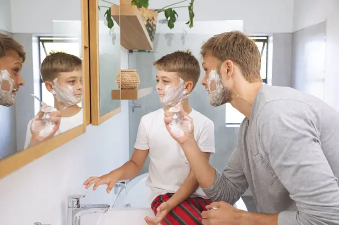 Caucasian man at home with his son together, in bathroom, shaving with shaving cream on faces, looking at mirror. Social distancing during Covid 19 Coronavirus quarantine lockdown.