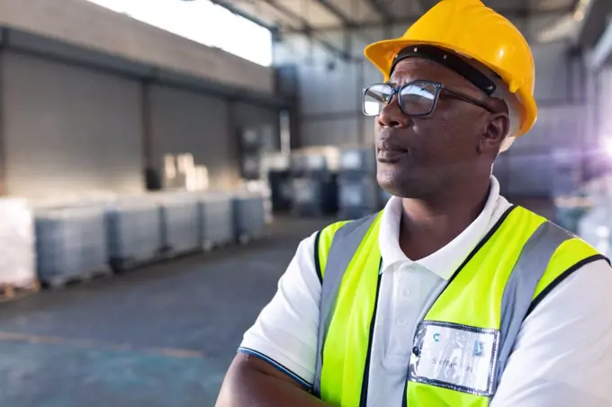 Portrait of Thoughtful African-american male staff in hardhat and reflective jacket standing with arms crossed in warehouse. This is a freight transportation and distribution warehouse. Industrial and industrial workers concept