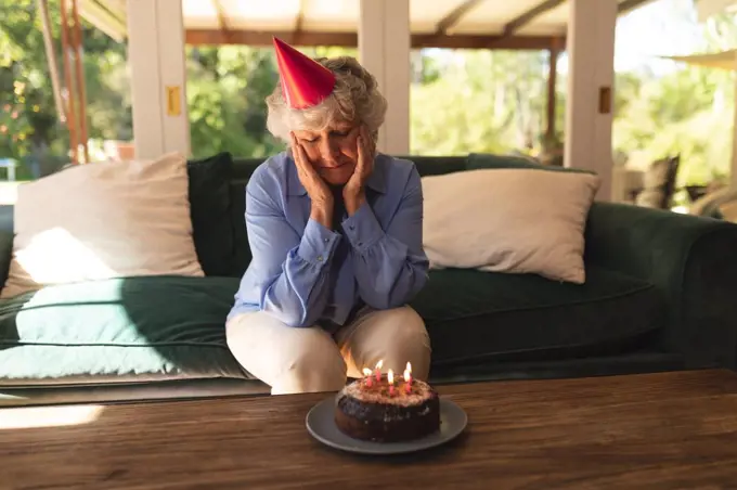 Senior caucasian woman spending time at home celebrating a birthday, wearing party hat and looking at cake. self isolation at home during coronavirus covid 19 quarantine lockdown.