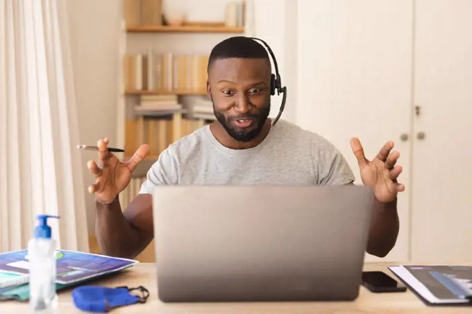 African american man using phone headset while having a video chat on laptop while working from home. social distancing during covid 19 coronavirus quarantine lockdown.
